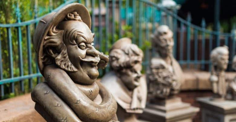 A close-up of whimsical stone busts with exaggerated facial features, displayed outdoors in a row—each one seeming to express its own guest pet peeves—framed by a decorative iron fence and lush greenery in the background.