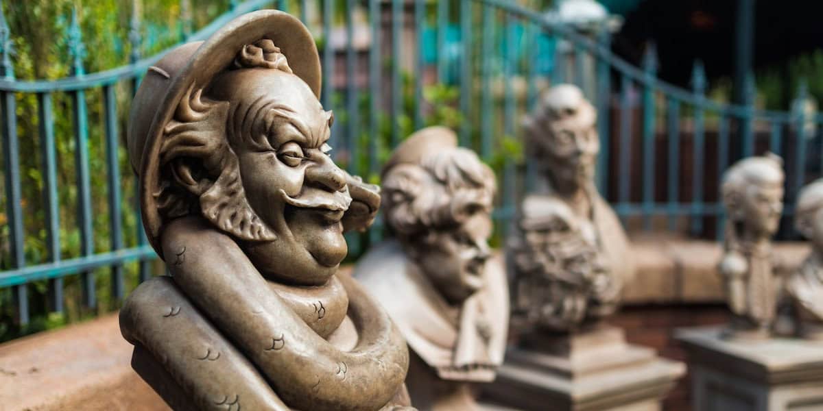 A close-up of whimsical stone busts with exaggerated facial features, displayed outdoors in a row—each one seeming to express its own guest pet peeves—framed by a decorative iron fence and lush greenery in the background.
