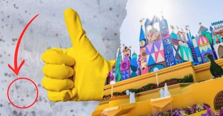 A gloved hand giving a thumbs-up is prominently displayed on the left side of the image. On the right side, there is a colorful and whimsical Disney building with various decorations and two small fountains in front, all under bright sunlight. A red arrow points to a small circled area on the left side.