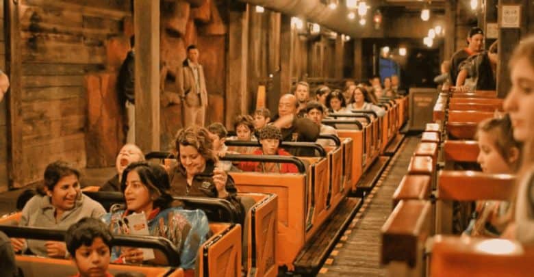 A group of people, including children and adults, are seated in a roller coaster train inside a dimly lit tunnel on the Big Thunder Mountain Railroad. Some are smiling and chatting, while a child in the front row is yawning. The atmosphere is lively and anticipatory.