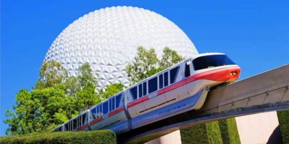 A monorail glides along a track in the foreground with the iconic geodesic sphere of epcot's spaceship earth in the background under a clear blue sky.