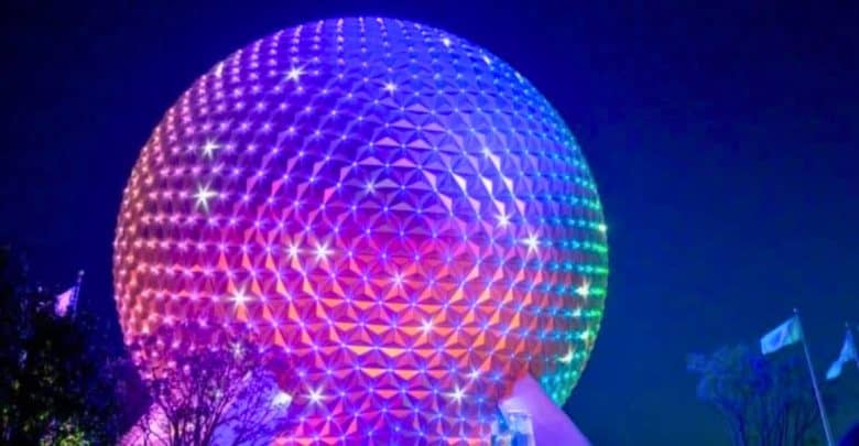 A large geodesic sphere, known as Spaceship Earth, is illuminated in vibrant rainbow colors against the night sky. Flags can be seen in the bottom right near trees silhouetted by the glow of the lights.