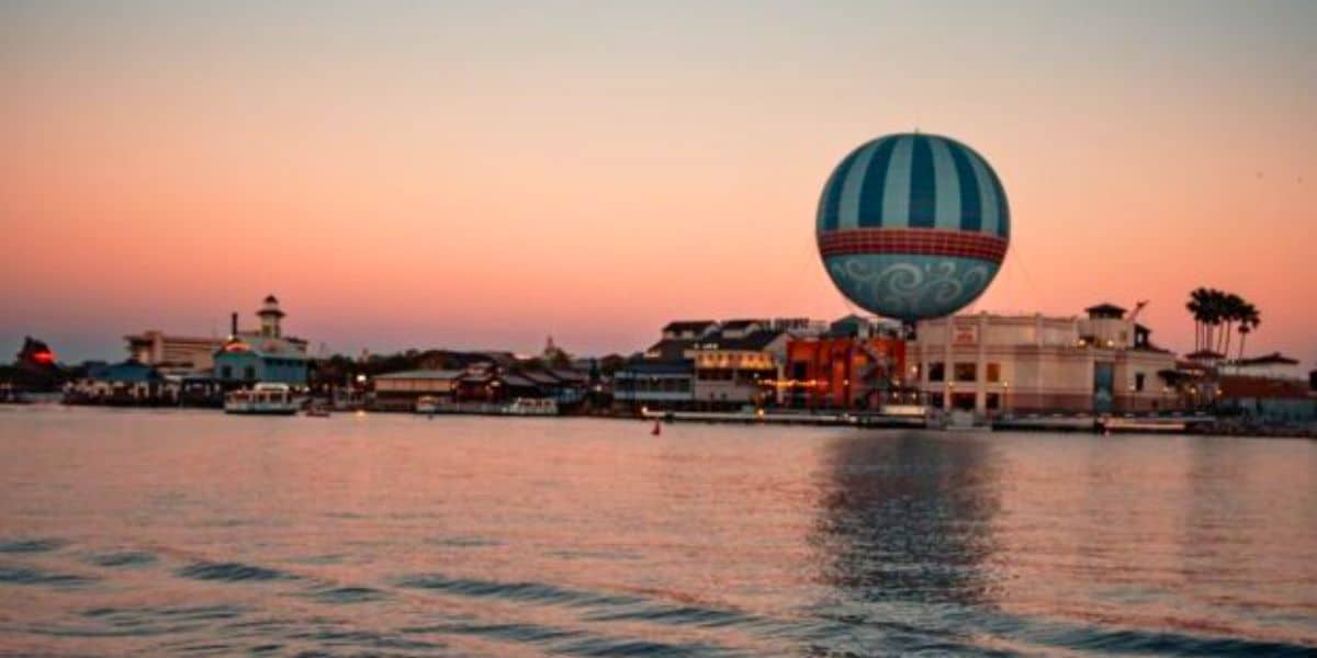 A large, colorful hot air balloona tethered at the waterfront of Disney Springs during sunset. The sky is pink and orange, and the calm waters reflect the buildings and the balloon. The shoreline features various buildings and palm trees in the background.