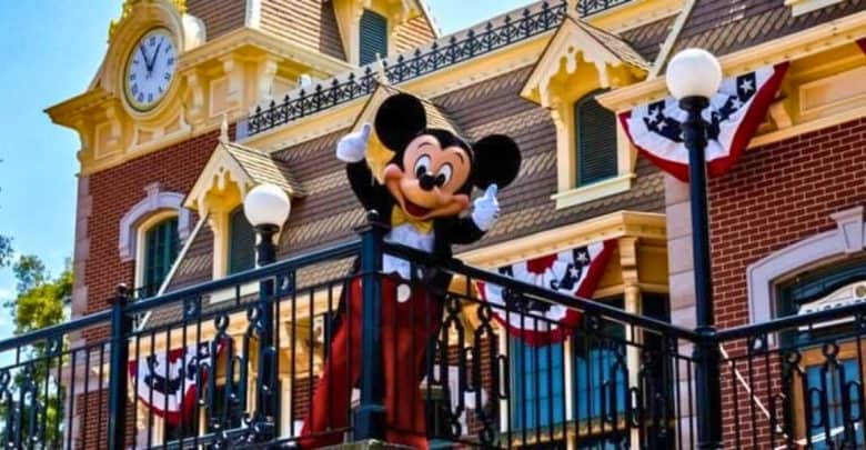 A person in a Mickey Mouse costume waves from a balcony at a theme park. The building behind them features a large clock and decorative elements, including red, white, and blue bunting. As the sky remains clear and blue, visitors are reminded to check for any Disney lost items.