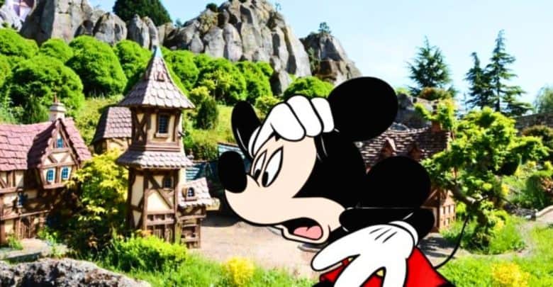 Image of mickey mouse superimposed on a scenic miniature village with detailed cottages and lush greenery surrounded by rocky mountains under a clear sky.