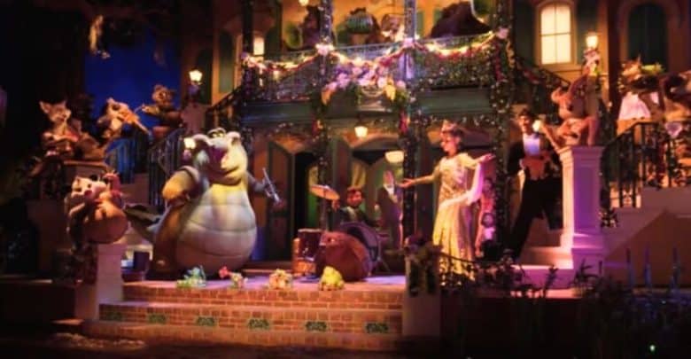 A vibrant scene featuring various colorful characters from Disney's "The Princess and the Frog" is set in front of a lively, decorated New Orleans-style building. The characters are in mid-performance, with musicians playing instruments and others dancing and singing, the finale of Tiana’s Bayou Adventure.