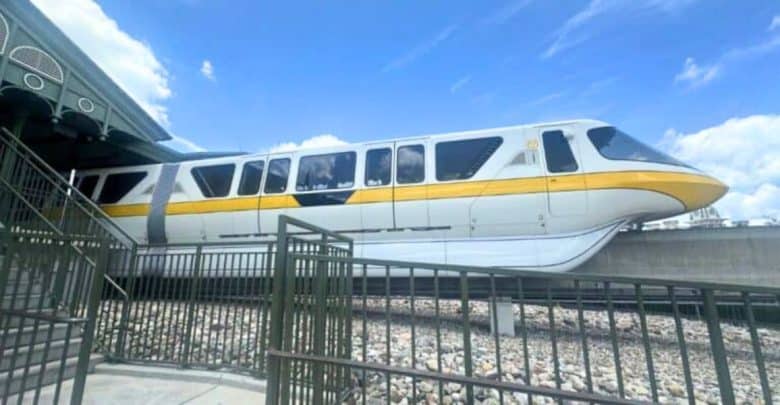 A Disney monorail train in yellow and white colors passing by on an elevated track at a Disney theme park, with a partly cloudy blue sky in the background and a metal fence in the foreground.