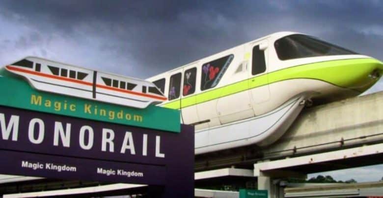 A green and white monorail glides along an elevated track under a cloudy sky. In the foreground, a purple sign with the words "Magic Kingdom Monorail" and an illustration of the monorail indicates the entrance to the transportation system.