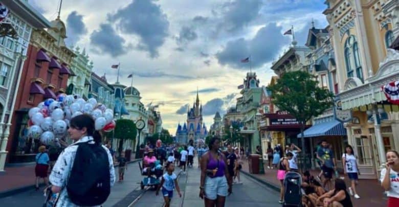 A bustling street scene at a theme park featuring people walking down the main street with the iconic castle in the background. Balloons, decorated shopfronts, and flags add to the festive atmosphere. The sky is partly cloudy, capturing a lively and vibrant moment.
