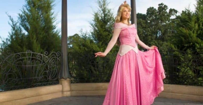 A woman dressed as a princess, wearing a pink ball gown, smiling and posing on a gazebo with green trees in the background.