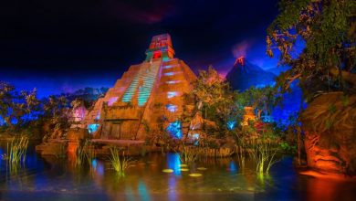 A vibrant night scene at EPCOT showcases an ancient pyramid, illuminated with colorful lights. Surrounding the pyramid are lush trees and vegetation, reflected in the calm water below. In the background, a volcano emits a soft red glow, enhancing the mystical atmosphere perfect for an evening of drinking and revelry.