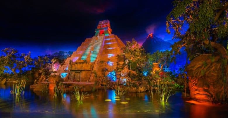 A vibrant night scene at EPCOT showcases an ancient pyramid, illuminated with colorful lights. Surrounding the pyramid are lush trees and vegetation, reflected in the calm water below. In the background, a volcano emits a soft red glow, enhancing the mystical atmosphere perfect for an evening of drinking and revelry.