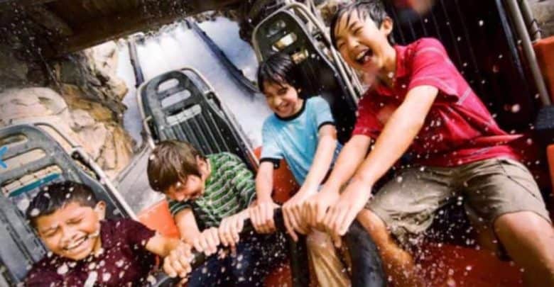 Four children are seated in a row on a log ride at a Disney attraction, holding on tightly and smiling as water splashes around them. They appear to be enjoying the thrill and excitement of the ride. The background shows part of the rocky structure and water slide.