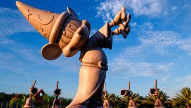 A stone statue of Mickey Mouse dressed as the Sorcerer's Apprentice, wearing a pointed hat and robe, with one arm raised, against a blue sky with scattered clouds. In the background, similar statues and palm trees are visible—one of the magical things to do at night at Disney without a ticket.