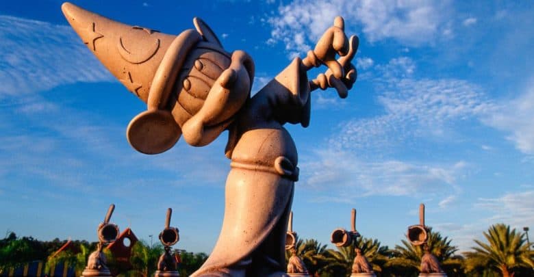 A stone statue of Mickey Mouse dressed as the Sorcerer's Apprentice, wearing a pointed hat and robe, with one arm raised, against a blue sky with scattered clouds. In the background, similar statues and palm trees are visible—one of the magical things to do at night at Disney without a ticket.
