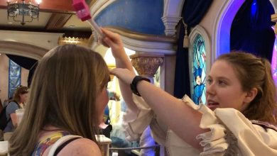 A woman in a costume playfully taps another woman's head with a wooden mallet inside a themed, colorful room at Walt Disney World, featuring arched doorways and a decorative stained-glass window. Other people are seen in the background enjoying free things to do around the park.