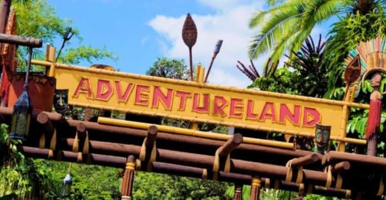 A colorful entrance sign to Adventureland featuring decorative masks and bamboo accents against a backdrop of lush tropical greenery and blue skies.