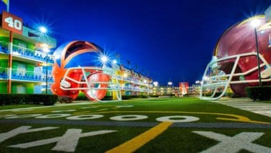 A football-themed outdoor area at night, featuring two giant helmet structures facing each other. The ground is painted with yard lines and hash marks resembling a football field. Surrounded by the lively ambiance of Disney World Resorts, the buildings are lit up, creating a vibrant atmosphere.