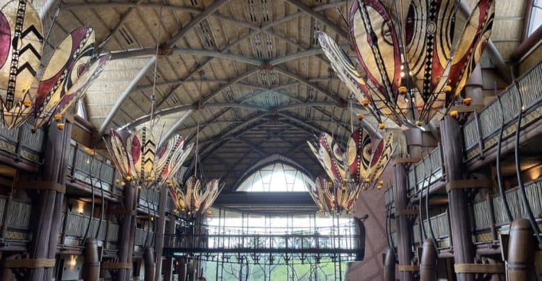The interior of a large, lodge-style building at Disney World Resorts features tall ceilings with wooden beams and decorative, leaf-shaped chandeliers. Sunlight streams through large, arched windows at the far end, illuminating the spacious and intricately designed hall.