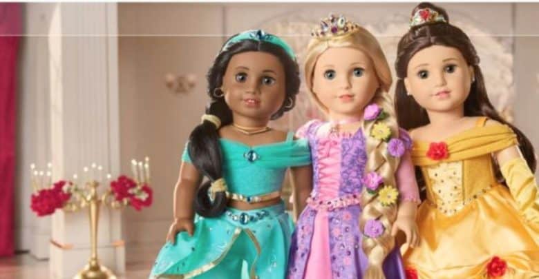Three dolls dressed as princesses are standing together in a lavish hall with red drapery and candles. From left to right, one American Girl doll wears a teal outfit and jeweled headband, another in a pink and purple gown with a long braid, and the third in a yellow Disney-inspired gown.