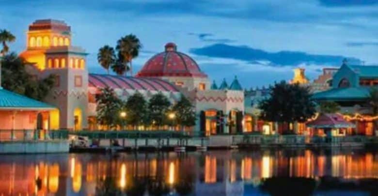 A scenic view of a waterfront area at dusk, featuring brightly lit, Mediterranean-style buildings with domed and tower-like structures. Palm trees dot the landscape, and the still water below reflects the colorful lights and architecture.
