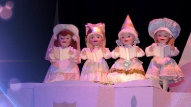 Four puppets dressed in elaborate white costumes stand in a row, holding songbooks. Each puppet wears a different headpiece: a bonnet, a cat-eared hat, a party hat, and a bonnet with ruffles. They are posed as if singing together, with soft purple lighting.