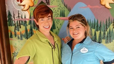 Two people stand smiling in front of a colorful, cartoon-like forest mural. One is dressed as Peter Pan in green; the other wears a blue shirt with a nametag, showcasing Disney World Cast Member Jobs.