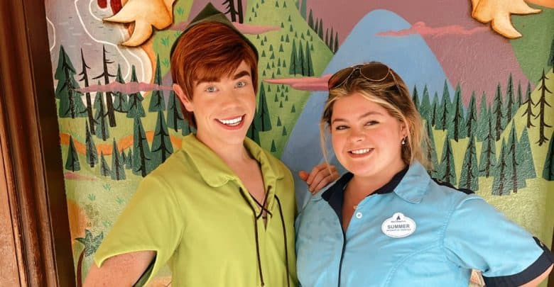 Two people stand smiling in front of a colorful, cartoon-like forest mural. One is dressed as Peter Pan in green; the other wears a blue shirt with a nametag, showcasing Disney World Cast Member Jobs.