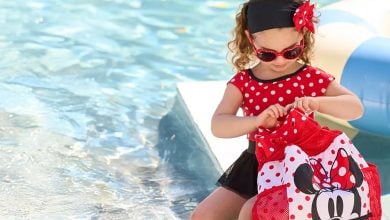 A young girl in a red polka-dot dress, red sunglasses, and a black headband sits by a pool, opening her Walt Disney World Essentials Minnie Mouse backpack. There is a pool float in the background.
