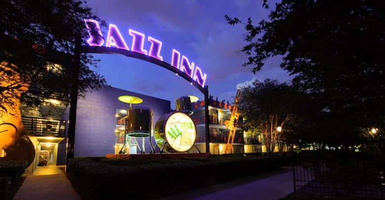 A brightly lit entrance to a building at night features a large, glowing purple "Jazz Inn" sign and oversized musical instrument decorations—hallmarks of the Best Disney Value Resort—surrounded by trees and illuminated pathways.
