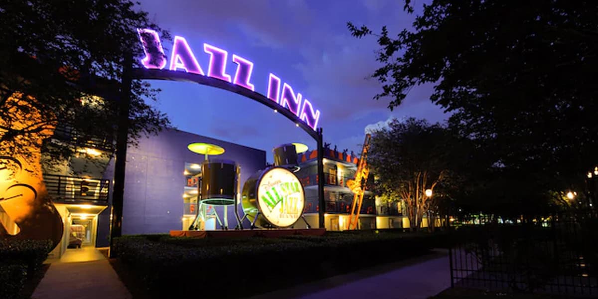 A brightly lit entrance to a building at night features a large, glowing purple "Jazz Inn" sign and oversized musical instrument decorations—hallmarks of the Best Disney Value Resort—surrounded by trees and illuminated pathways.
