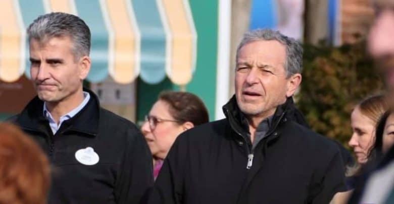 A group of people standing outdoors with a colorful, striped awning in the background. The two men in the foreground are dressed warmly in black coats. The man on the left is wearing a name tag. Other individuals are visible but out of focus in the background.