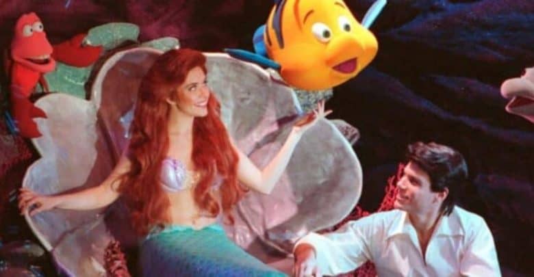 A scene from "The Little Mermaid" shows Ariel sitting in a large seashell, looking up at Flounder, who is floating above her hand. Sebastian the crab is beside her. Eric, wearing a white shirt, is sitting nearby, engaged in the moment.