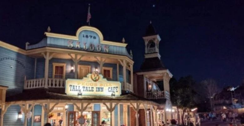 A nighttime scene featuring a well-lit Old West-style saloon with an American flag on the roof. The saloon's sign reads "Pecos Bill Tall Tale Inn & Cafe." Decorative wooden architecture and tower add to the historical Western vibe. People are walking nearby.