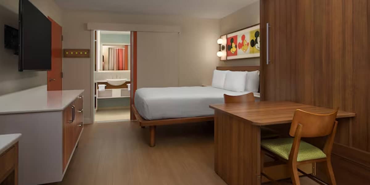 Modern hotel room with a queen bed, wooden furniture, a desk with chair, flat-screen TV, and an open-door bathroom. Colorful Mickey Mouse artwork above the bed highlights this space at the Best Disney Value Resort.
