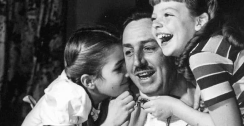 A black and white photo of a man smiling and wearing a mustache is surrounded by two young girls. One girl is whispering into his ear while the other is laughing and has her arm around him. All are sharing a joyful moment together.