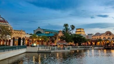 A scenic view of Coronado Springs at dusk reveals a waterfront village with colorful, architecturally diverse buildings adorned with domes and arches. Lines of palm trees stand gracefully as the water mirrors both the structures and the clear evening sky.