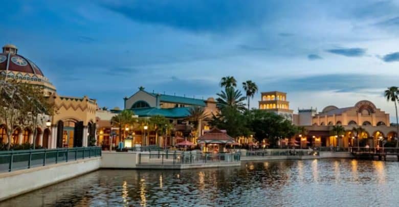 A scenic view of Coronado Springs at dusk reveals a waterfront village with colorful, architecturally diverse buildings adorned with domes and arches. Lines of palm trees stand gracefully as the water mirrors both the structures and the clear evening sky.