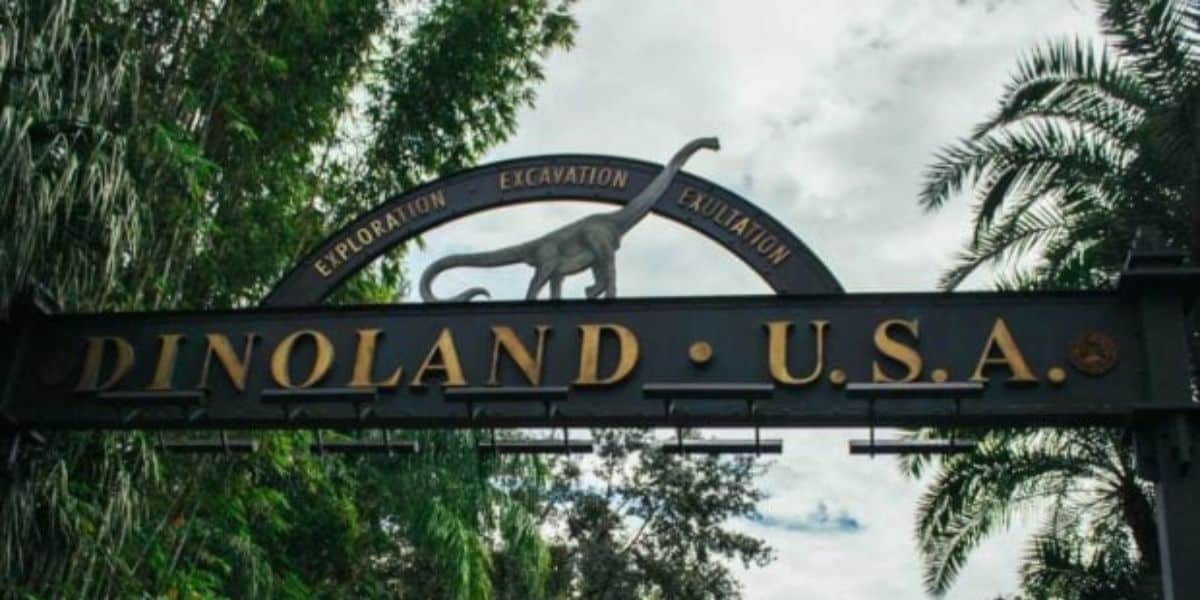 Entrance archway to Dinoland U.S.A. with a dinosaur silhouette and words "Exploration", "Excavation", and "Exultation" above. Surrounded by lush trees and a partly cloudy sky.