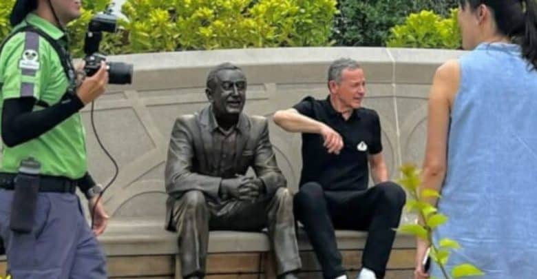 A man in a black shirt sits on a bench next to a bronze statue of a seated man. A woman stands nearby facing them, and another person wearing a green shirt and carrying a camera is on the left. Green bushes form the background.
