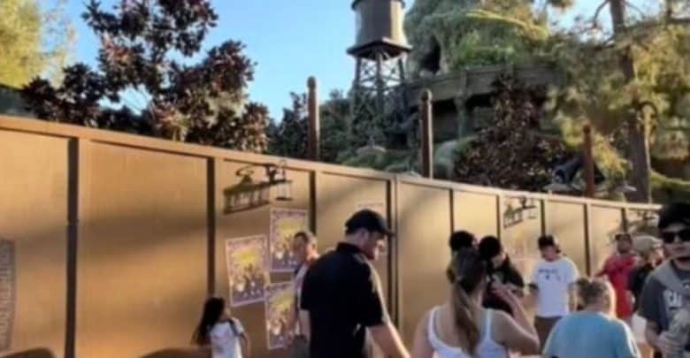 A group of people are gathered near a wooden fence with posters, in an outdoor area surrounded by trees and foliage. A water tower is visible in the background under a clear blue sky.