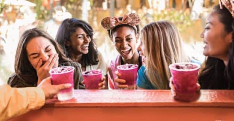 A group of five women smiling and laughing while holding purple frozen drinks. They appear to be having fun at an outdoor setting with greenery in the background. One woman's hand is visible, offering another drink.