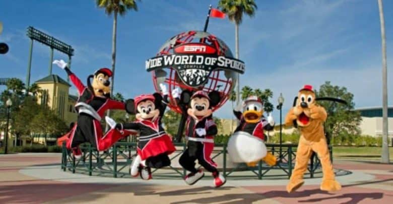 Characters in sports attire are jumping joyfully in front of the ESPN Wide World of Sports globe. The scene is outdoors, with palm trees and a clear sky in the background.