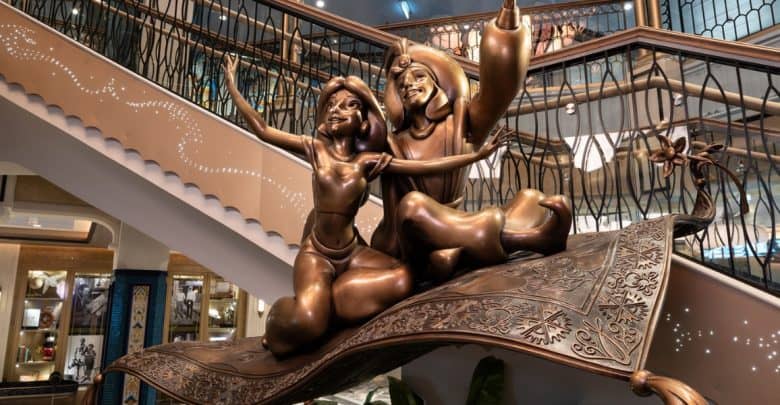Bronze statue of two animated characters on a flying carpet, situated under a staircase in an elegantly decorated interior space. The scene suggests a sense of joy and adventure.