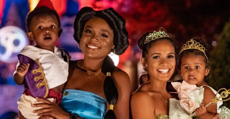 Two women and two young children dressed as Disney princesses and characters, smiling together at night with colorful lights and a castle in the background, all perfectly following the Disney dress code.