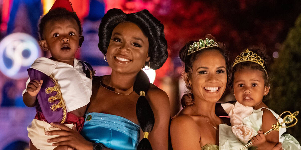 Two women and two young children dressed as Disney princesses and characters, smiling together at night with colorful lights and a castle in the background, all perfectly following the Disney dress code.
