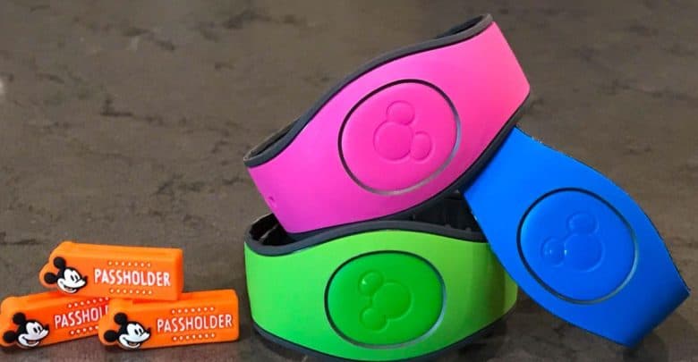 Three colorful wristbands—pink, blue, and green—are stacked together. Each has a Mickey Mouse logo. Two orange passholder tags with Mickey Mouse designs are placed beside them on a gray surface.