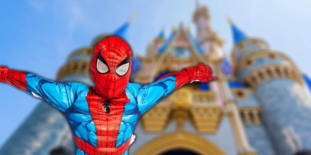 A kid in a Spider-Man costume, poses with arms outstretched in front of a blurred, colorful Disney castle on a sunny day.