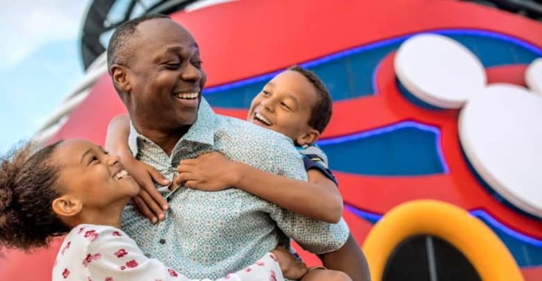 A smiling man joyfully holds two children, one in each arm. They are in front of a colorful structure with a large Mickey Mouse emblem, suggesting a Disney-themed location. The group looks happy and engaged in a lighthearted moment.