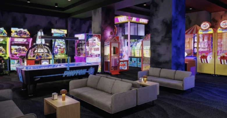 A vibrant arcade featuring neon-lit game machines, including racing games, claw machines, and ticket redemption games. In the foreground, there are sofas and tables with drinks, providing a lounge area for relaxation.